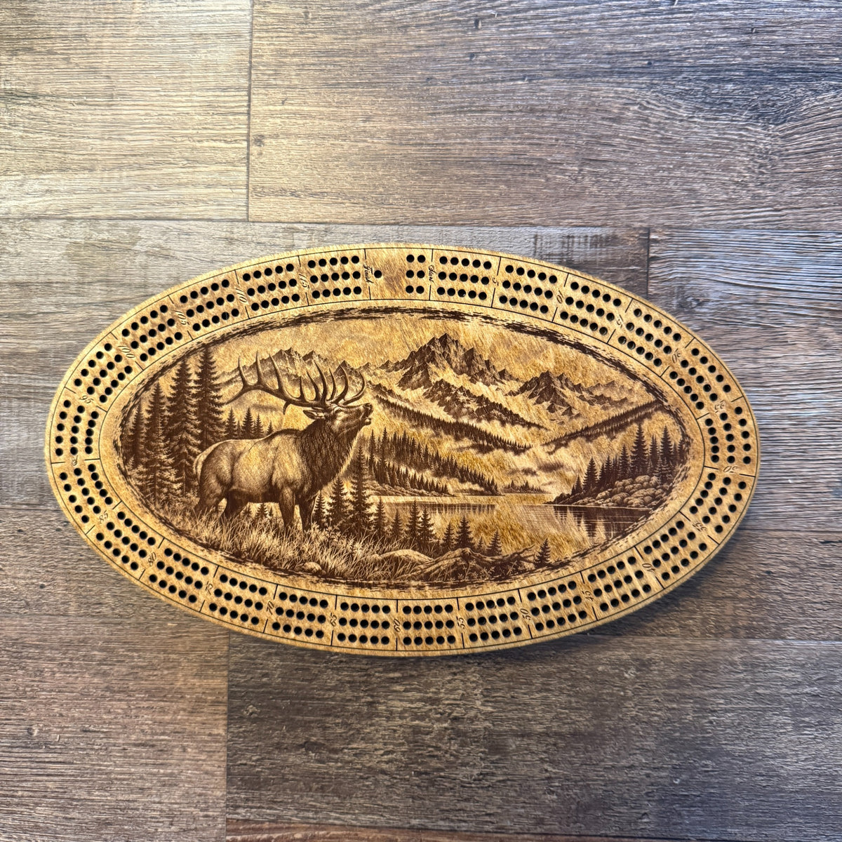 Handcrafted oval cribbage board featuring a bugling bull elk, a mountain landscape and a lake